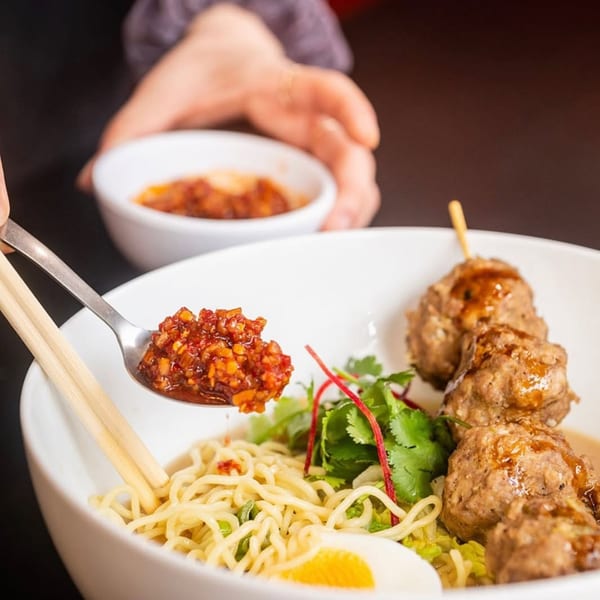 A hand adds chili oil to a bowl of Kameya Ramen with meatballs, noodles, and a soft-boiled egg.