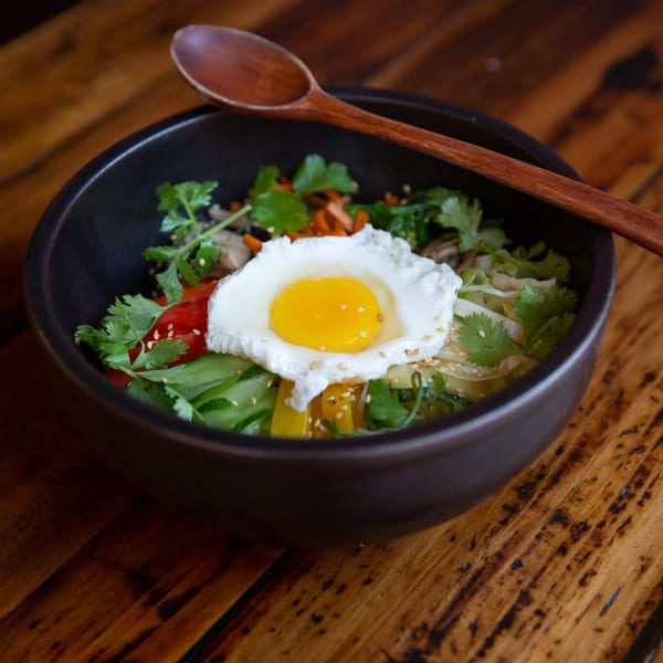 Kameya Ramen & Sushi's colorful bibimbap bowl with a fried egg and wooden spoon on a rustic table.