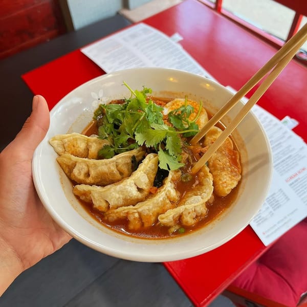 A hand holding a bowl of Kameya Ramen & Sushi's gyoza in broth with cilantro and chopsticks.